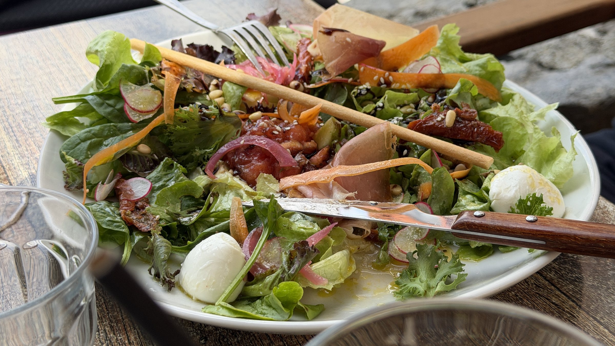 Salade italienne servie en terrasse : mesclun, jambon cru, billes de mozzarella, tomates séchées, pignons et gressins